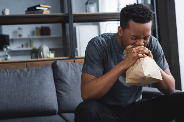 a man sitting on a couch blowing his nose into a paper bag